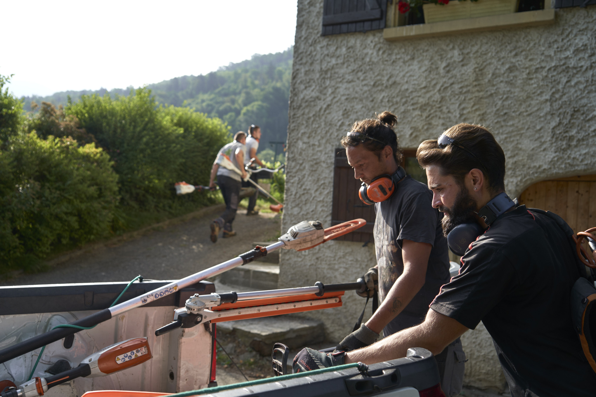 Двоє ландшафтних архітекторів вивантажують акумуляторні пристрої STIHL з автомобіля, на задньому плані — інші працівники 