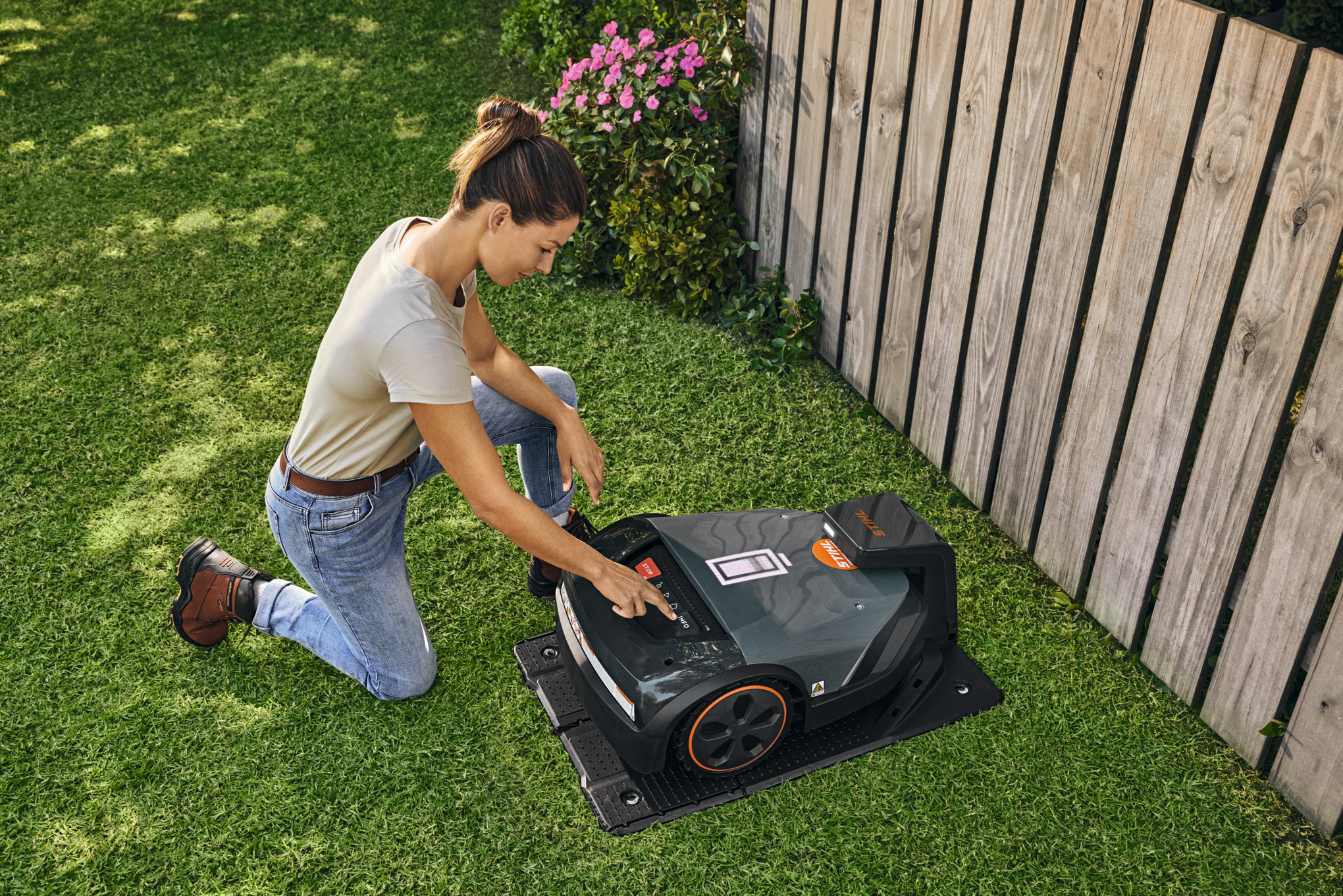 Газонокосарка-робот STIHL iMOW® на док-станції в саду, поки жінка щось налаштовує на газонокосарці-роботі.
