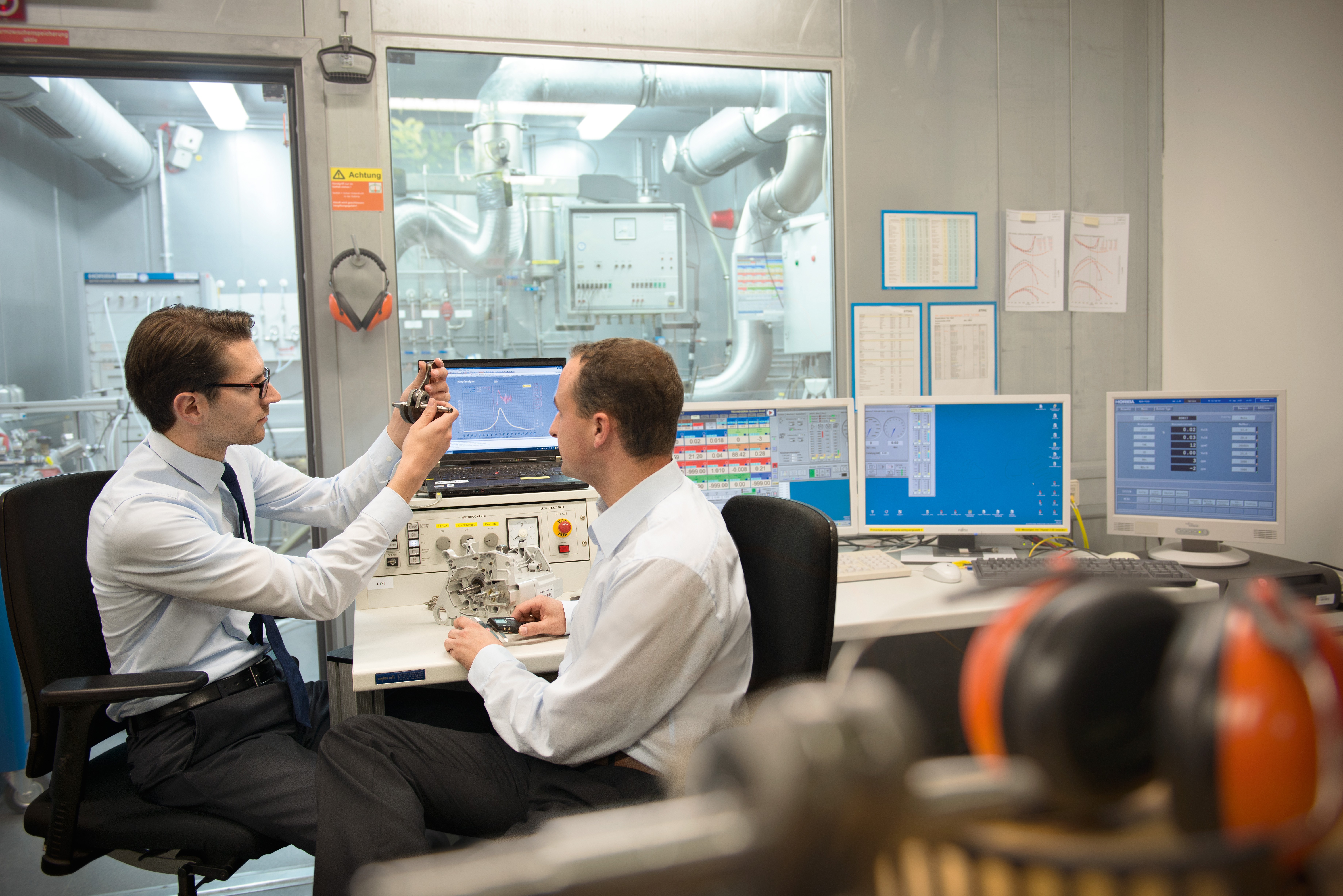 Zwei Männer sitzen in einem Labor an einem Schreibtisch mit mehreren Minitoren und begutachten Teil eines neuen Produktes
